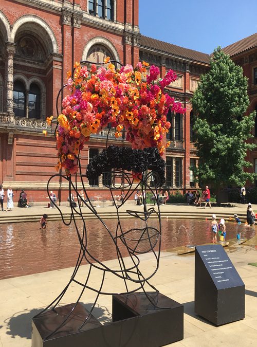 A hige black wire frame of a head with pink and orange flowers as the hair outside the V&A Museum