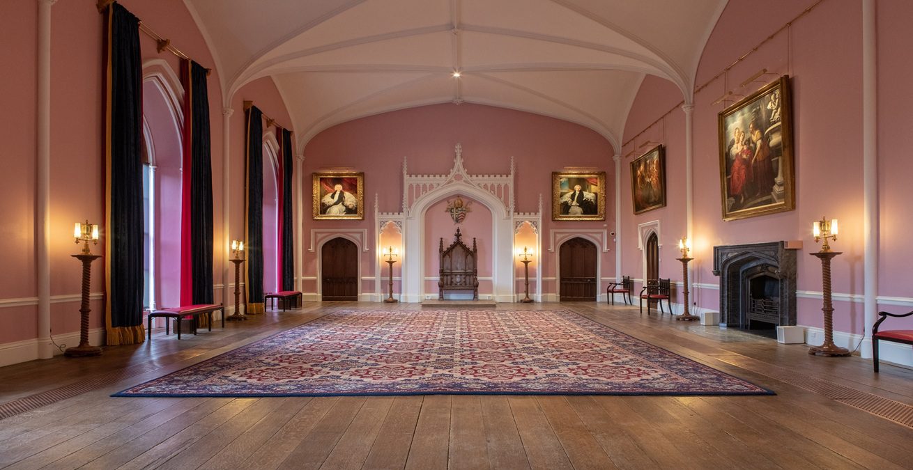 A pink painted room with high vaulted white ceiling, paintings of people on the walls and long dark red curtains at the windows in Auckland Castle