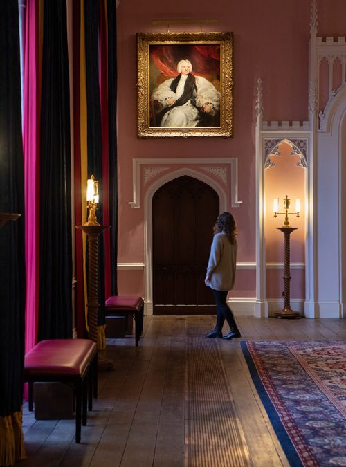A pale pink painted room in Auckland Castle with long red and black floor length curtains and a painting of a man in white on the back wall