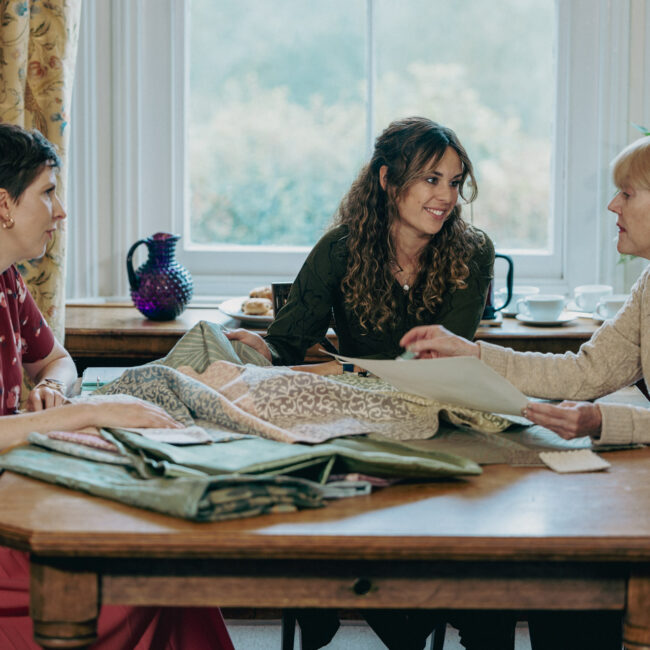 Two account managers talking through a project with a customer, in the Humphries Weaving Showroom. Sitting at a table with a pile of fabric and inspiration.