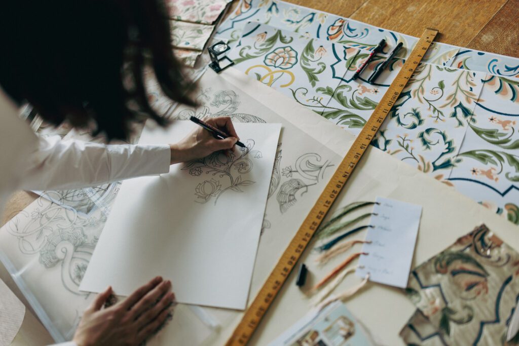 Woman leaning on table with printed design, hand-drawing and tracing elements, surrounded by pens, yarn tabs, a ruler and a pick glass
