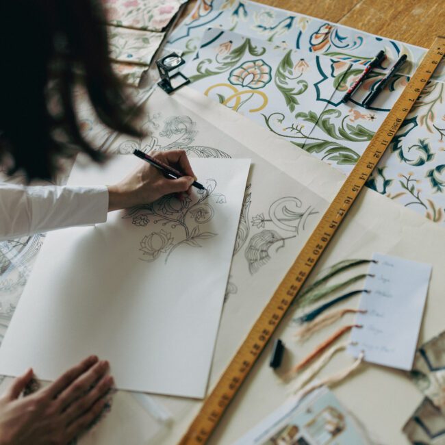 Woman leaning on table with printed design, hand-drawing and tracing elements, surrounded by pens, yarn tabs, a ruler and a pick glass