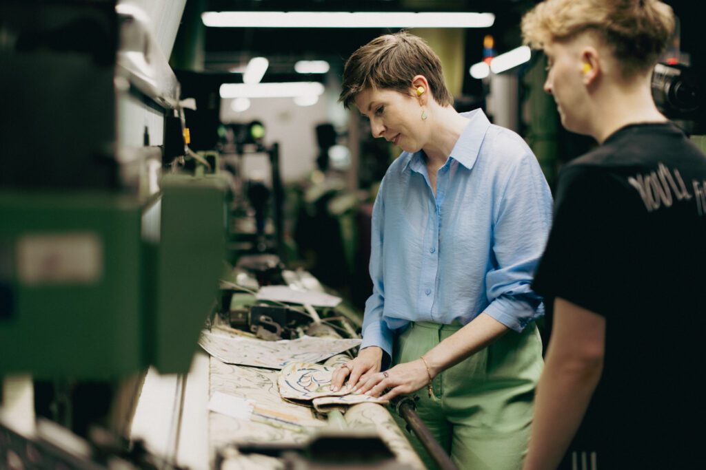 Account Manager Sarah Wall standing by a loom with a weaver, holding a piece of fabric and comparing it to the fabric on the loom