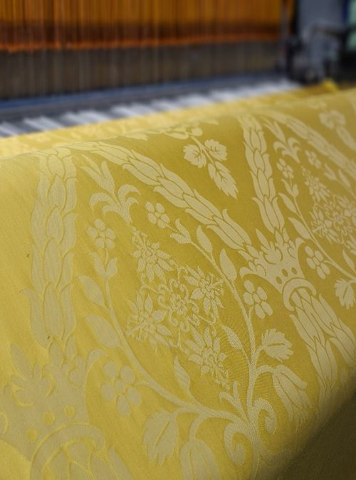 A close up image of Rosa Mundi weaving on a jacquard loom. The fabric has a gold background and beige gothic figure