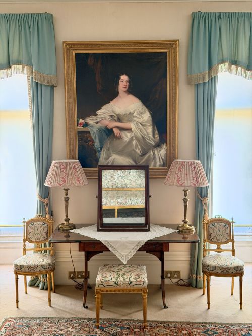 Elegant interior of the Mercia Room at Highclere Castle, featuring blue drapery, embroidered furnishings, and a framed textile display beneath a large portrait.